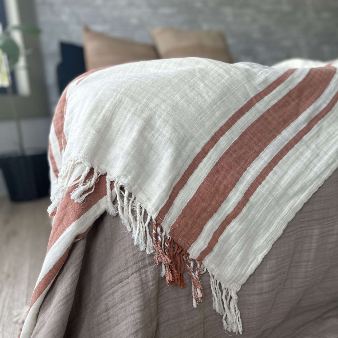 A textured cotton throw blanket draped over a bed, featuring alternating stripes of off-white and a muted terracotta rust color, finished with fringed tassels.