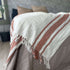 A textured cotton throw blanket draped over a bed, featuring alternating stripes of off-white and a muted terracotta rust color, finished with fringed tassels.